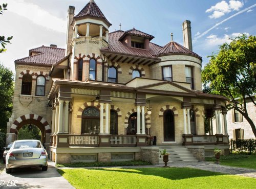 A large, ornate mansion with a turret, intricate brickwork, and a well-manicured lawn under a blue sky.