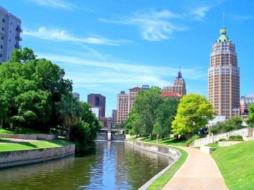 A scenic view of a river lined with trees, leading to a city skyline under a clear blue sky.