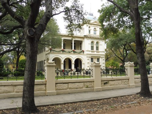 A historic white mansion with a decorative balcony, surrounded by trees and a wrought-iron fence.