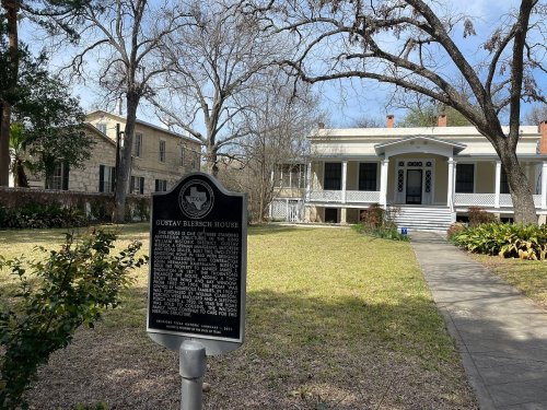 Historic Gustav Blersch House with a plaque in front, surrounded by trees and a well-maintained lawn.