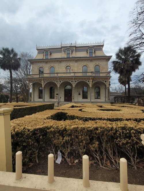 A grand, historic mansion with ornate architecture, surrounded by neatly trimmed hedges and palm trees under a cloudy sky.