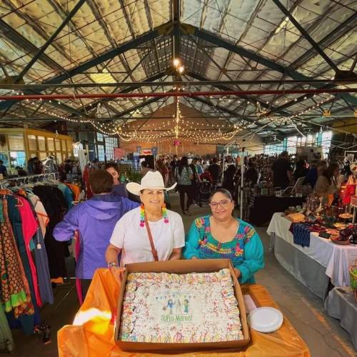 Two vendors smile while holding a large cake at a bustling market event, with colorful decorations and people in the background.