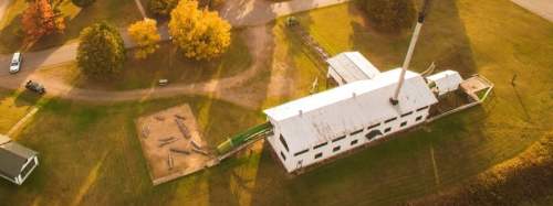Aerial view of a white building with a tall chimney, surrounded by trees and a playground area on a sunny day.