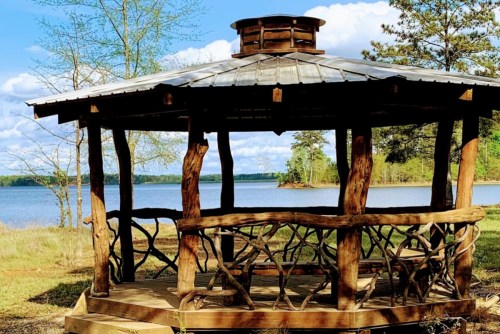 A wooden gazebo by a lake, surrounded by trees and a clear blue sky with fluffy clouds.