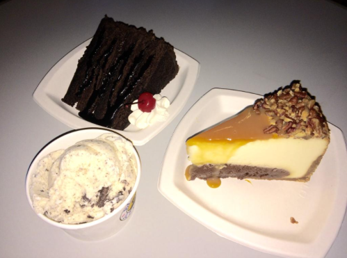 A plate with a slice of cheesecake topped with nuts, a piece of chocolate cake, and a cup of cookies and cream ice cream.