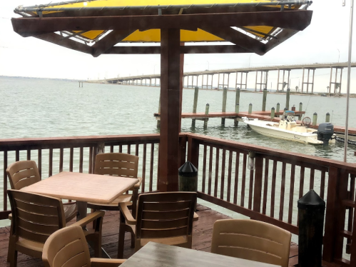 A waterfront view from a deck with wooden tables and chairs, featuring a boat and a bridge in the background.