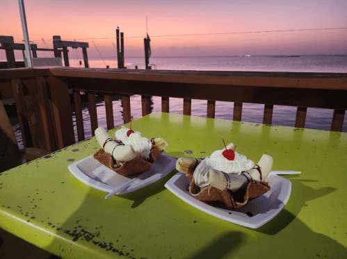 Two banana splits topped with whipped cream and cherries sit on a green table, with a sunset over the water in the background.