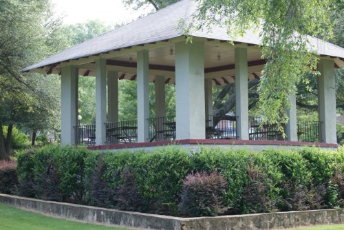 A green gazebo with columns surrounded by lush bushes in a park setting.