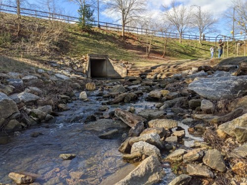 A rocky stream flows through a park, with a drainage pipe and trees in the background under a partly cloudy sky.