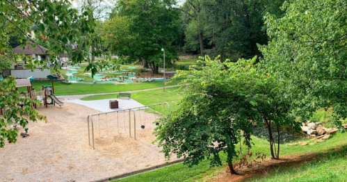 A playground with swings and a sandy area, surrounded by green trees and a grassy park.
