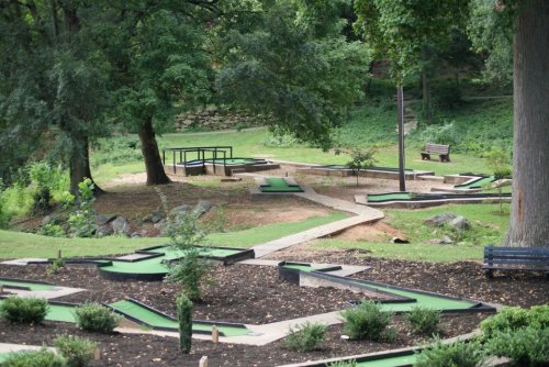A mini-golf course with winding paths, surrounded by trees and greenery, featuring benches and a picnic table.