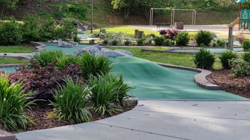 A mini-golf course with green turf, landscaped bushes, and a playground in the background.