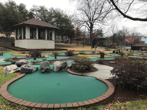 A mini-golf course with green turf, rocks, and a gazebo in a park setting, surrounded by trees and pathways.