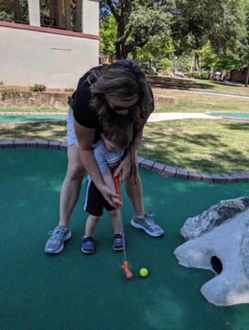 A woman helps a young child putt a golf ball on a mini-golf course, surrounded by greenery.