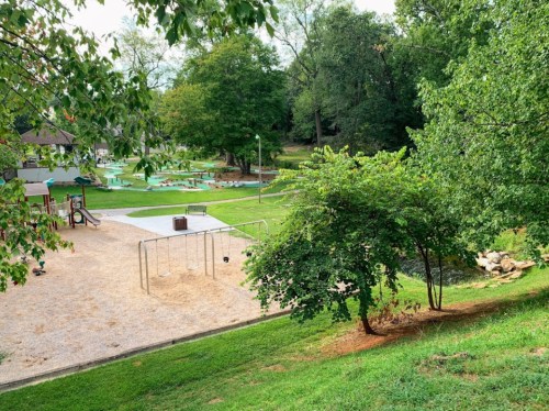 A playground with swings and a grassy area, surrounded by trees and a small creek in the background.