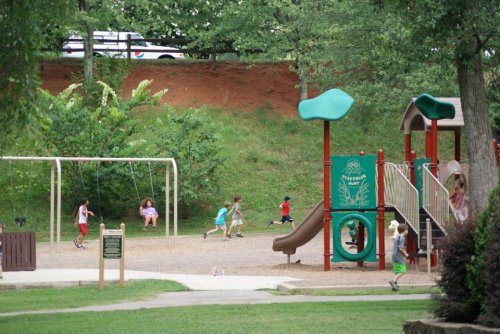 Children playing in a park with swings, a slide, and a playground structure surrounded by greenery.
