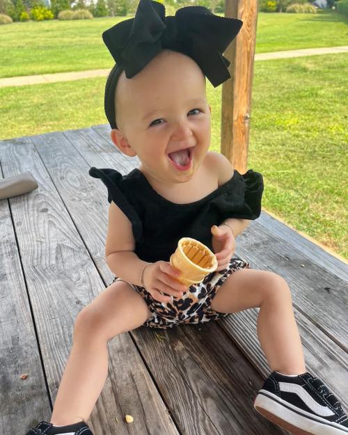 A joyful toddler with a big bow enjoys an ice cream cone while sitting on a wooden deck.