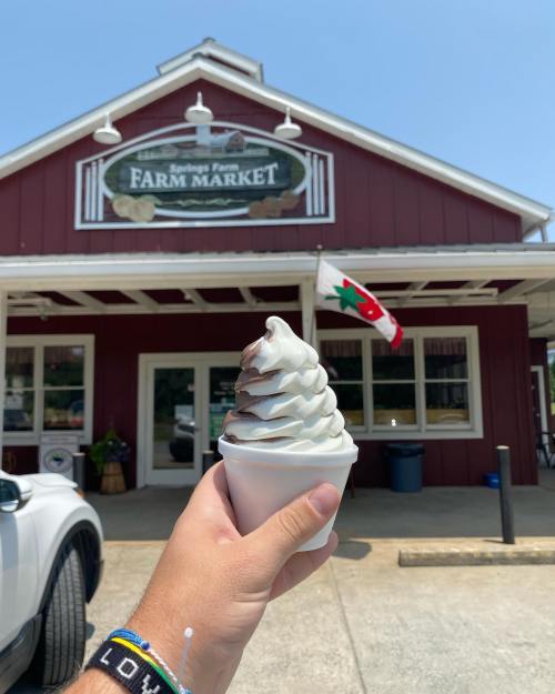 A hand holds a soft serve ice cream cone in front of a red farm market building with a flag.