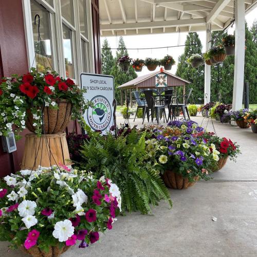 Colorful flower baskets and pots adorn a porch area, with tables and chairs set up nearby under a covered structure.