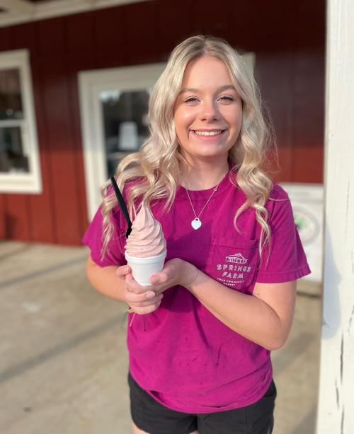 A smiling young woman in a pink shirt holds a soft serve ice cream cone outside a building.