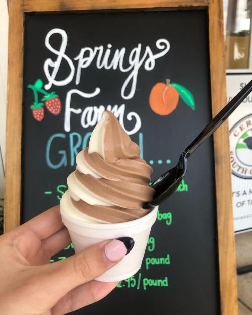 A hand holding a cup of swirled chocolate and vanilla soft serve ice cream in front of a colorful sign.