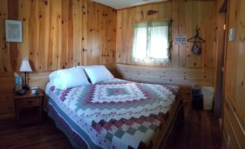 Cozy wooden bedroom with a quilted bedspread, window, lamp, and a small wastebasket. Warm, rustic decor.