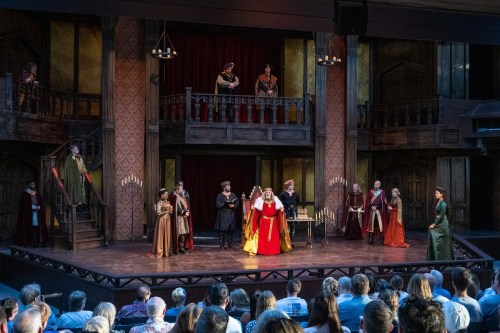 A theatrical performance with actors in period costumes on stage, surrounded by an audience in a historic setting.