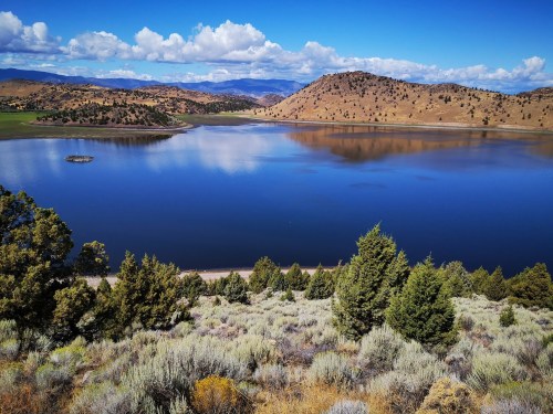 A serene lake surrounded by hills and trees, reflecting clouds and blue sky in the calm water.