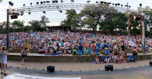 A large crowd of people seated on colorful blankets at an outdoor concert venue, with a stage in the background.