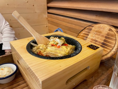 A wooden bowl with a sizzling dish topped with vegetables and a wooden spoon, served on a wooden tray.