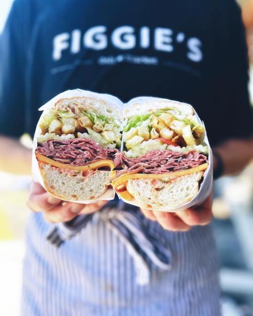 A person holds a large sandwich cut in half, showcasing layers of meat, cheese, and fresh vegetables.