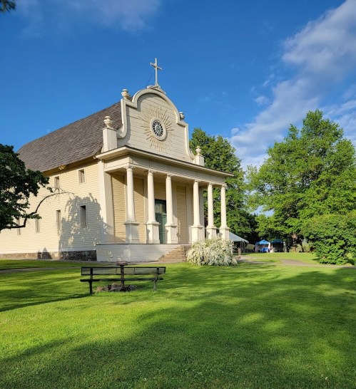 A church with a cross atop, featuring columns and a round window, surrounded by green grass and trees under a blue sky.