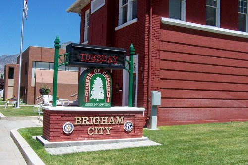 Sign for the Brigham City Chamber of Commerce, featuring visitor information, with a red brick building in the background.