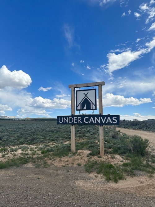 Sign for "Under Canvas" with a tent icon, set against a blue sky with clouds and a dirt road.