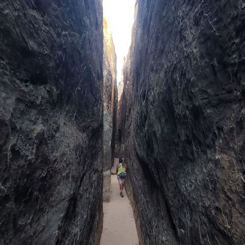 A narrow canyon with steep rock walls, featuring a person walking along a sandy path between the cliffs.