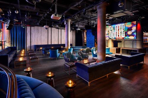 A stylish lounge with blue velvet seating, low tables, and a vibrant screen displaying colorful visuals.