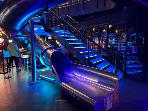 A metallic slide illuminated with blue lights, next to a staircase in a modern indoor space.