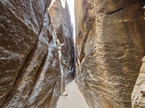 Narrow canyon walls of rugged rock with a sandy path in between, illuminated by sunlight above.