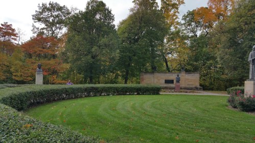 A landscaped park with manicured grass, bushes, and two busts surrounded by trees in autumn colors.