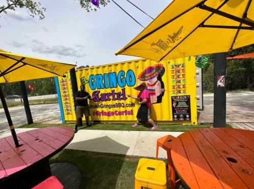 Two people stand by a colorful food truck with a yellow container, surrounded by bright outdoor seating and umbrellas.