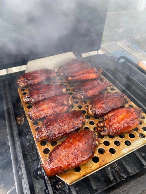 Grilled chicken wings on a perforated tray, surrounded by smoke, cooking on a barbecue grill.