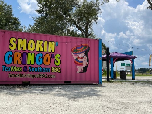 A colorful food truck with a pink exterior, featuring "Smokin' Gringo's TexMex & Southern BBQ" signage and a shaded seating area.