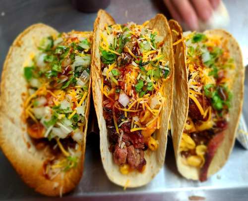Three loaded tacos filled with meat, cheese, onions, and cilantro, served on a metal surface.
