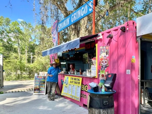 A colorful food truck named "Smokin' Gringos" with a vendor in a cowboy hat, surrounded by trees.