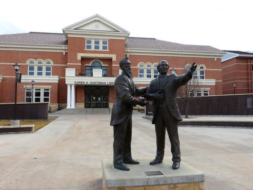 Statue of two men shaking hands in front of the Karen W. Huntsman Library, with a brick building in the background.