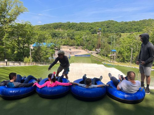 A group of people sitting in blue inflatable tubes on a grassy slope, preparing for a tubing activity in a scenic outdoor setting.