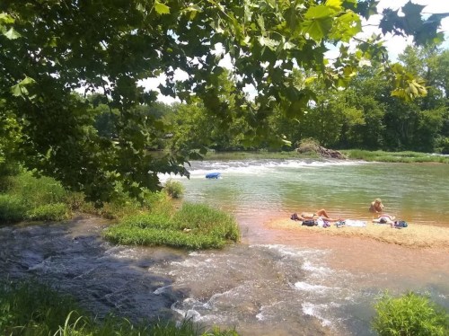 A serene river scene with people relaxing on the shore, surrounded by lush greenery and gentle water flow.