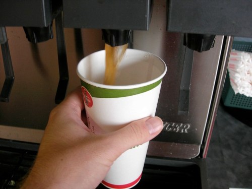 A hand holds a cup under a beverage dispenser as a drink is being poured into it.