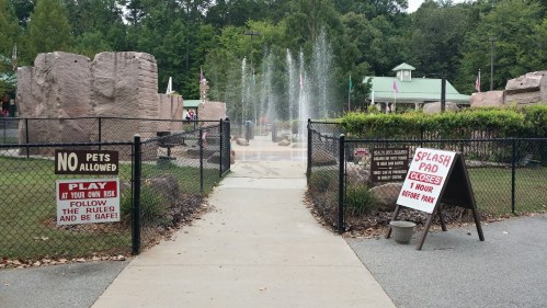 A pathway leads to a splash pad area with signs about safety and pet restrictions, surrounded by greenery and fountains.