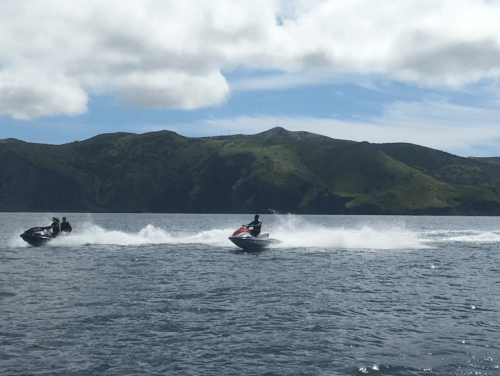 Two jet skis race across a calm sea, with green hills and a cloudy sky in the background.